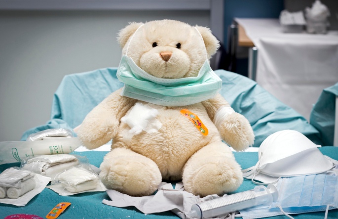 Cream colored teddy bear sitting on a medical exam table with sterile bandages and other medical supplies, with a bandaid on its chest and wearing a paper mask.